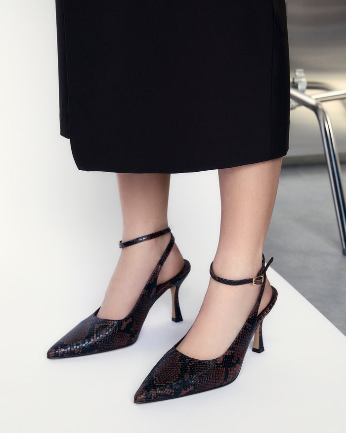 Black high-heeled shoes with snake print worn by a person in a black skirt.