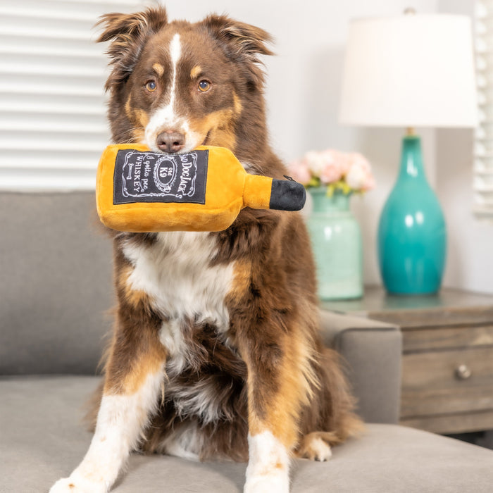 Dog holding a yellow bottle-shaped plush toy featuring Bad Dog Jack's Whiskey.