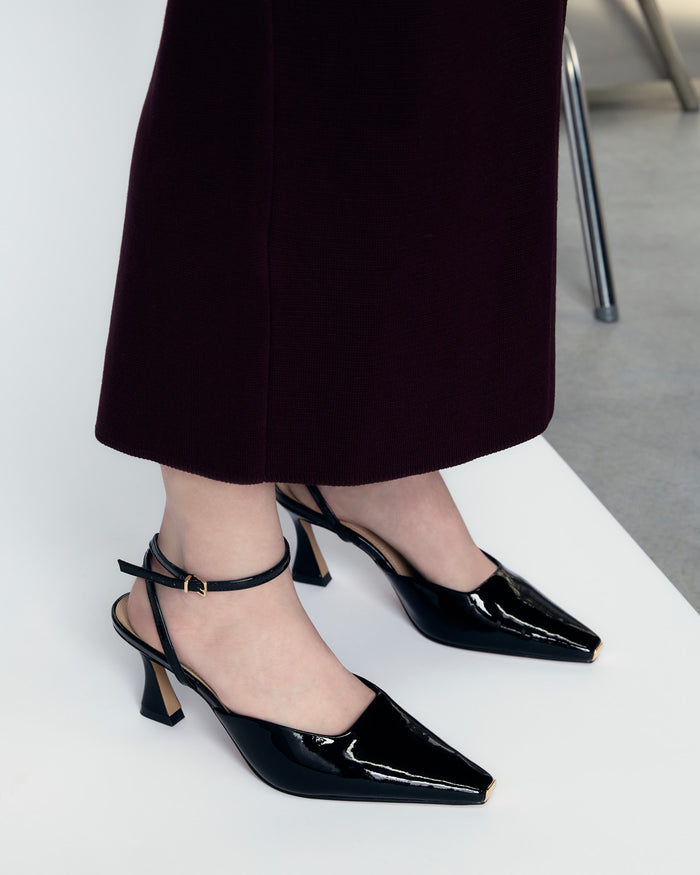 Black patent leather shoes worn with a dark skirt on a white floor.
