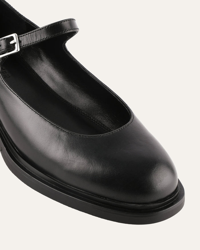 Black leather flat shoe with a strap on a light gray background